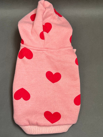 UNA SUDADERA CON CAPUCHA EN COLOR ROSA Y ESTAMPADO DE CORAZONES ROJO. MANGA LARGA, SE METE TIPO JERSEY, SIN CIERRES NI BOTONES. ES CALIENTITA PORQUE POR DENTRO ES UNA FELPITA DE PIEL DE DURAZNO.